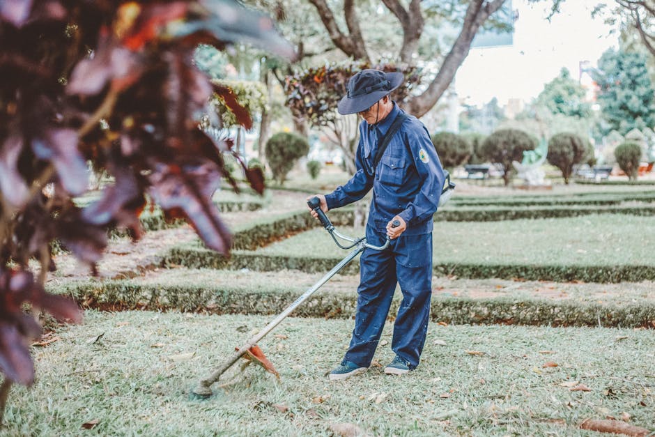 A gardener dressed in a dark blue uniform and a wide-brimmed hat is using a grass trimmer to maintain a neatly trimmed lawn in a well-organized park setting. The soil near the edge of the grassy area