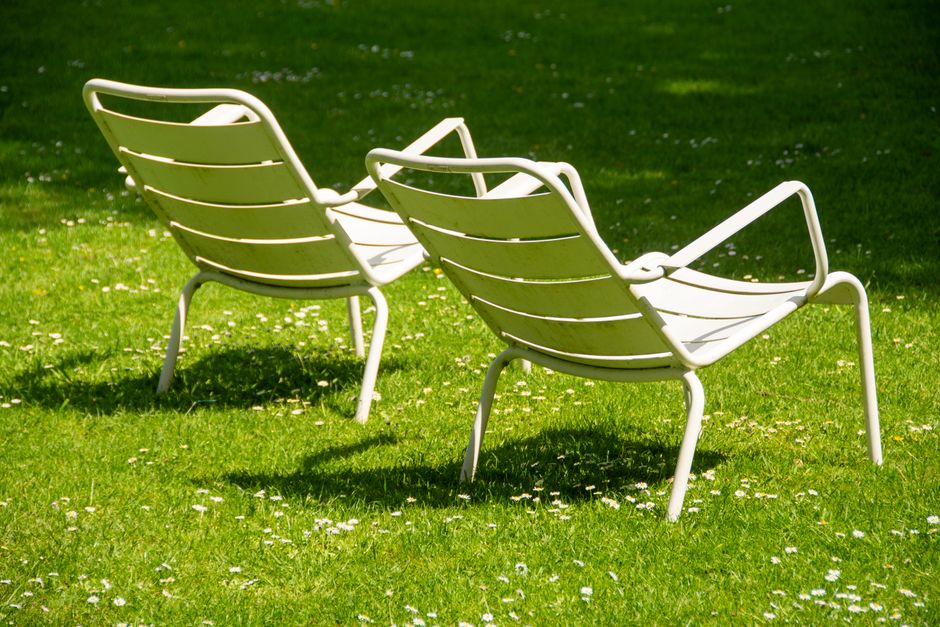 Two white metal garden chairs with curved slatted seats and backs are positioned on a vibrant green lawn, with one slightly behind the other. The lawn features small white flower petals scattered acro